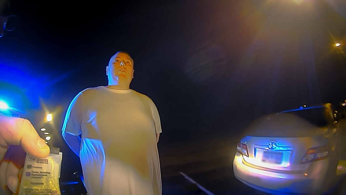 A screenshot from a police body camera video showing a suspect standing in handcuffs and an officer's hand holding a field drug test kit in the foreground | Police body camera footage