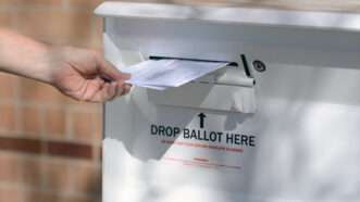 A hand places a ballot into a ballot drop-box.