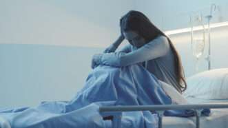 Stock photo of a woman with long hair, sitting in a hospital bed and looking upset.