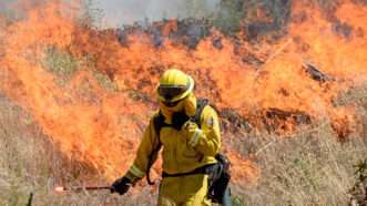 Fireman spraying brush while fire burns in the background. | Will Lester/ZUMA Press/Newscom