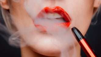 Woman with red lipstick has vapor coming out of her open mouth, with a vape pen near her lips.