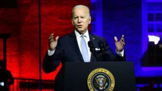 Joe Biden speaks in front of a podium with the presidential seal with red and blue lighting in the background