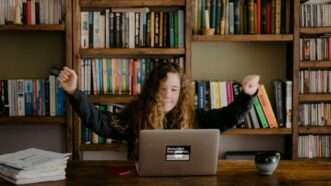 Child raising her arms with a laptop in front of her and wall of bookshelves behind her.