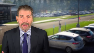 John Stossel stands in front of a row of cars