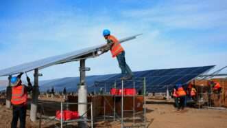 People in orange vests work on a row of solar energy panels. | FEATURECHINA/Newscom