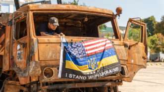 A man in a baseball hat stands in a rusted vehicle hanging a combination U.S. and Ukrainian flag that says "I stand with Ukraine."