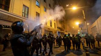 A riot policeman fires tear gas over the heads of a line of riot police ahead of him, in a nighttime urban environment. | Lucas Aguayo Araos/dpa/picture-alliance/Newscom