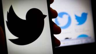 black Twitter logo on screen of phone being held by shadowy hand