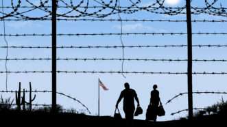 shadowed figures of two people holding bags walk toward a flying American flag as seen through a hole in a barbed wire fence | Photo 186902785 © Robert Goebel | Dreamstime.com