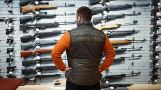A man in an orange shirt and a vest stands with his back to the camera in front of a wall of long guns, for sale and on display. | Nomadsoul1 | Dreamstime.com