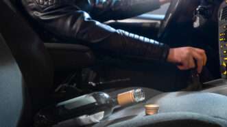 A man prepares to shift his car into gear as an empty wine bottle rests on the passenger seat.