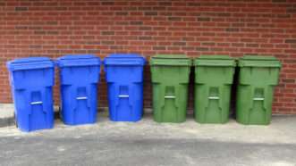 Six recycling bins, three blue and three green, lined up in a row against a brick wall | Rosamund Parkinson | Dreamstime.com