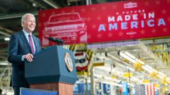 President Joe Biden, grinning, stands behind a lectern bearing the presidential seal. He is in a Detroit automobile factory with a giant red banner in the background that says "A FUTURE MADE IN AMERICA" | Adam Schultz/ZUMA Press/Newscom