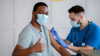 A man in a mask gives a thumbs up to the camera while a man in scrubs and a mask injects him with something