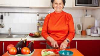 A smiling older woman cutting produce in a kitchen.