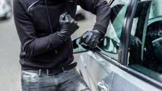 A thief wearing black gloves and a balaclava breaks into a car with a screwdrier. | Djedzura | Dreamstime.com