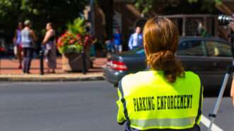 An officer with a neon PARKING ENFORCEMENT vest stands with back to camera, surveying a busy sidewalk scene. | Georgesheldon | Dreamstime.com