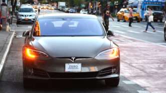 A Tesla sits in traffic, with a front-facing license plate that says "ZERO EMISSIONS" | Adrian825 | Dreamstime.com
