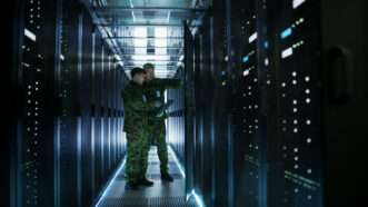 Two men in military uniforms stand between rows of data servers.