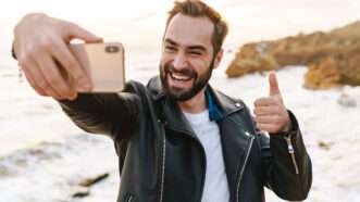 A man in a leather jacket walking along the beach takes a selfie on his phone. | Vadymvdrobot | Dreamstime.com