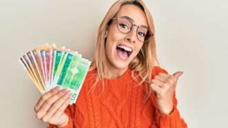 Smiling blonde women gives a thumbs up and holds a handful of Norwegian kroner, fanned out. | Aaron Amat | Dreamstime.com