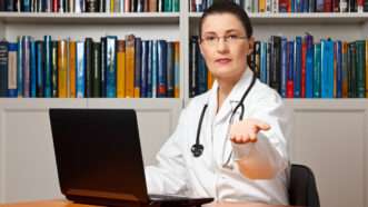 Female doctor sitting in front of a bookcase and behind a laptop with her hand sticking out, as if to ask for money.