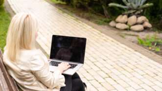 A blonde woman sits on a park bench on a laptop. | Andrew Angelov | Dreamstime.com