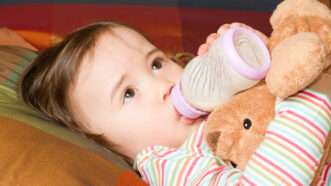 A baby girl with a stuffed animal drinks milk from a bottle. | Vitmark | Dreamstime.com