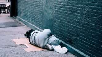 A homeless person sleeps on the street next to a brick wall | Photo by Jon Tyson on Unsplash