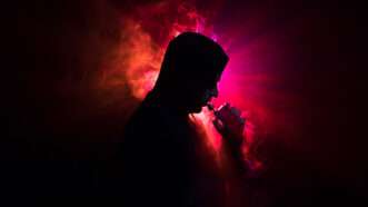 A shadowed figure of a man vapes with a red and pink glowing background behind the figure | Photo 112959091 © Ilkin Guliyev | Dreamstime.com