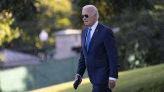 President Joe Biden wearing a suit and sunglasses walks past a background of green grass and green trees. | Chris Kleponis - Pool via CNP/CNP / Polaris/Newscom