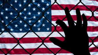 A hand grabs at a chain link fence with an American flag on the other side, symbolizing the struggle to immigrate to America. | Bradley Greeff | Dreamstime.com