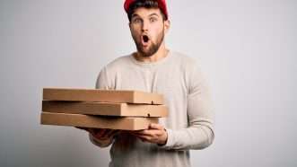 A man holds a stack of pizza boxes against a gray background | Photo 226993235 © Aaron Amat | Dreamstime.com
