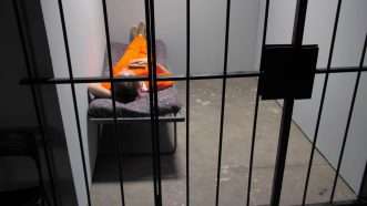 A single prisoner in an orange jumpsuit lies on a cot in a prison cell. | Ilya Ginzburg | Dreamstime.com