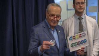 Sen. Chuck Schumer holds a container of Zyn nicotine pouches in his right hand and a piece of paper with other images of Zyn containers in his left hand. A man wearing a white coat and a tie stands behind him. | Ron Adar / M10s / MEGA / Newscom/RAAST/Newscom