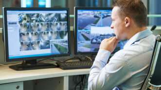 A man in a white Oxford shirt sits, monitoring screens that display footage from surveillance cameras. | Dmitry Kalinovsky | Dreamstime.com