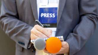 A journalist with a PRESS badge holds two microphones and writes in a notebook | Wellphotos | Dreamstime.com