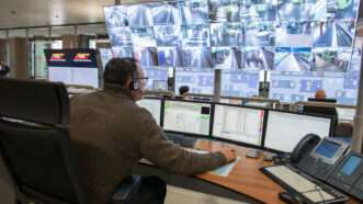 A man in an olive sweater sits in front of a bank of surveillance monitors. | Viappy | Dreamstime.com