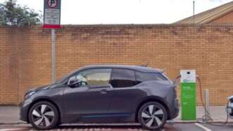 A hatchback electric vehicle charges at a public charger adjacent to a low brick building. | Mrnovel | Dreamstime.com