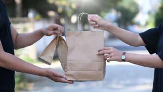 A delivery driver hands off a paper bag of takeout food to a customer. | Prathan Chorruangsak | Dreamstime.com