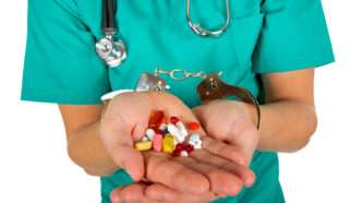 Close-up of a doctor's hands holdings pills, with his wrists handcuffed. Doctor is wearing green scrubs and a stethoscope around his neck. | DPST/Newscom