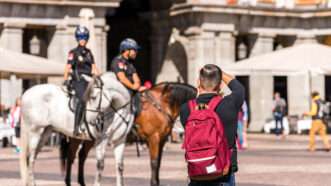 A photographer with a red backpack, in the foreground, takes pictures of police officers on horseback, in the background. | Gerold Grotelueschen | Dreamstime.com
