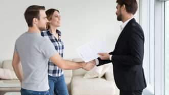 A realtor or landlord shakes hands with a young couple in a bright, sparsely-furnished apartment, while holding a rental agreement. | Fizkes | Dreamstime.com