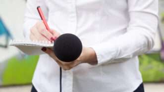 A reporter takes notes with a pad and pen while holding a microphone. | Wellphotos | Dreamstime.com