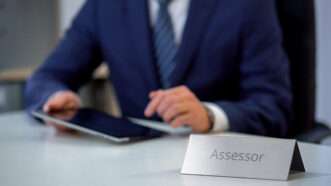 An unseen man in a business suit types on a tablet, at a desk with a nameplate that reads "Assessor" | Motortion | Dreamstime.com