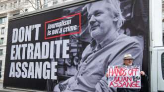 Man holding a sign stands in front of a billboard that reads, "Don't Extradite Assange" | Photo 175384998 © John Gomez | Dreamstime.com