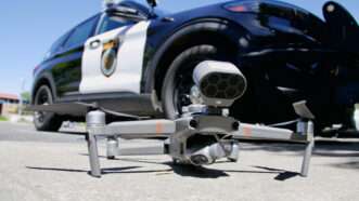 A drone with a camera attached sits on the ground, with a police car in the background. | Wirestock | Dreamstime.com