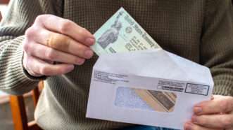 A man in a ribbed green sweater opens an envelope and takes out a Treasury check for COVID-19 pandemic stimulus. | Susan Sheldon | Dreamstime.com