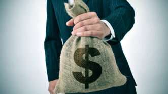 A man in a suit holds a burlap sack with a dollar sign on it | Photo 40229146 © Juan Moyano | Dreamstime.com