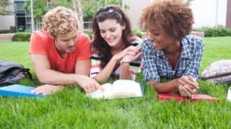 Three diverse coed college students lay on the grass, looking at a book. | Pkchai | Dreamstime.com